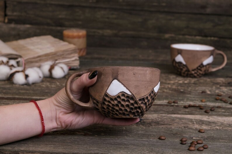 Może przedstawiać: Brązowy kubek ceramiczny z białym wzorem g&oacute;r. Kubek jest fakturowany i ma uchwyt. Kubek jest trzymany przez rękę z czerwoną bransoletką.