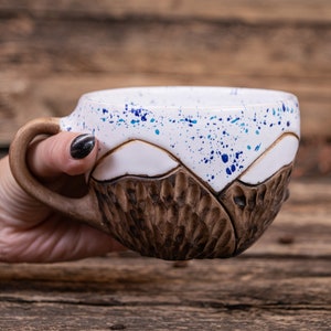 Może przedstawiać: Ceramiczny kubek z brązowym i białym wzorem g&oacute;rskim. Kubek ma niebieskie plamki na białej części. Kubek jest trzymany przez rękę.