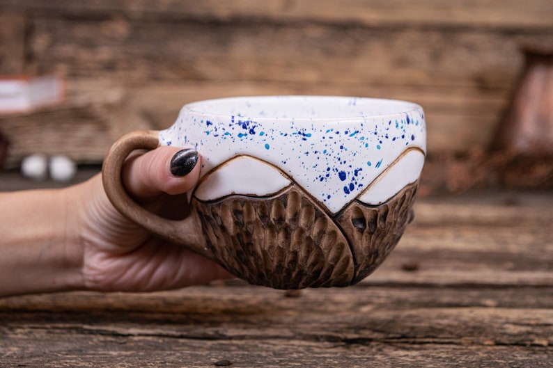 Może przedstawiać: Ceramiczny kubek z brązowym i białym wzorem g&oacute;rskim. Kubek ma niebieskie plamki na białej części. Kubek jest trzymany przez rękę.