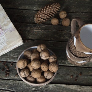 May include: A rustic wooden table with a bowl of walnuts, a pine cone, and two brown ceramic mugs with white accents. The table is also decorated with coffee beans.