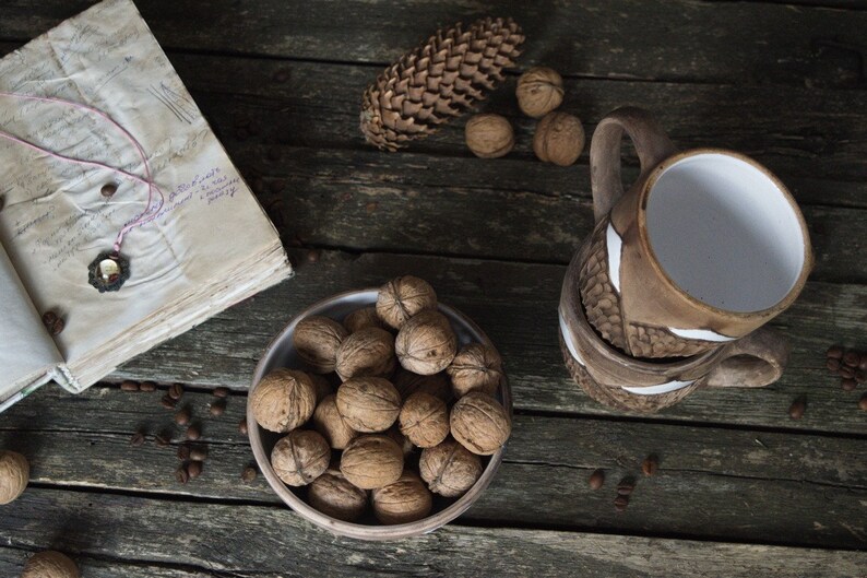 May include: A rustic wooden table with a bowl of walnuts, a pine cone, and two brown ceramic mugs with white accents. The table is also decorated with coffee beans.