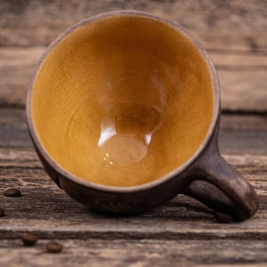 May include: A brown ceramic mug with a yellow glaze inside. The mug is sitting on a wooden surface.