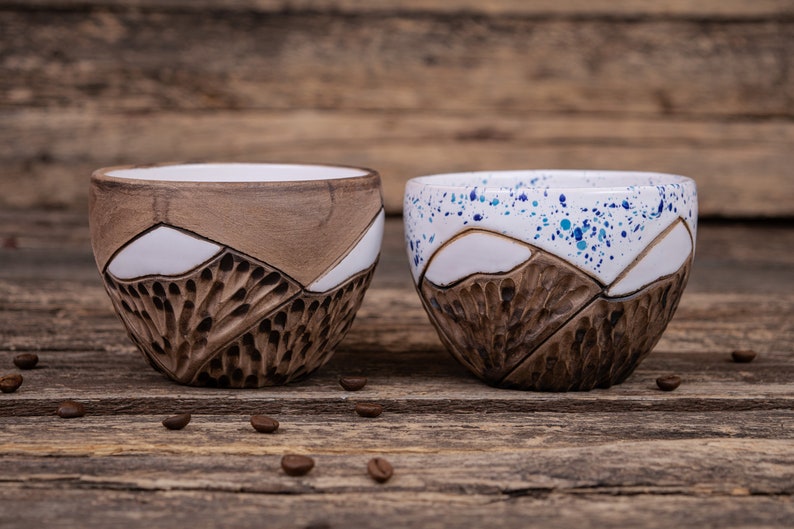 Może przedstawiać: Dwa ceramiczne kubki z wzorem g&oacute;r. Jeden kubek ma białą glazurę z niebieskimi plamkami, a drugi brązową glazurę. Oba kubki mają fakturowany wz&oacute;r g&oacute;r.