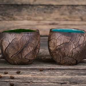 May include: Two brown ceramic mugs with leaf-like texture and green and blue glaze on the inside. The mugs are sitting on a wooden surface.