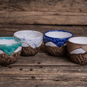 Może przedstawiać: Cztery ceramiczne kubki z wzorem g&oacute;rskiego pasma. Kubki są w r&oacute;żnych kolorach: niebieskim, białym, brązowym i zielonym. Kubki stoją na drewnianej powierzchni.