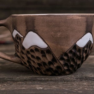 Może przedstawiać: Brązowy kubek ceramiczny z wzorem g&oacute;rskiego pasma. G&oacute;ry są białe, a kubek ma fakturowaną powierzchnię.