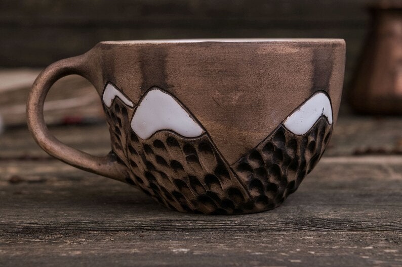 Może przedstawiać: Brązowy kubek ceramiczny z wzorem g&oacute;rskiego pasma. G&oacute;ry są białe, a kubek ma fakturowaną powierzchnię.