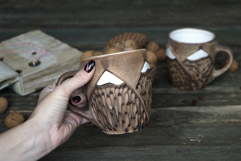 May include: Two brown ceramic mugs with white mountain designs. The mugs have a textured surface and are handmade.