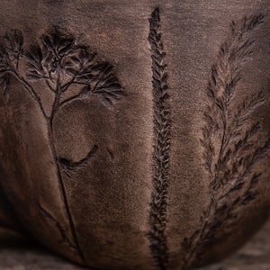 May include: A dark brown ceramic mug with a textured surface. The mug has three plant designs embossed into the surface.