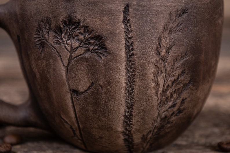 May include: A dark brown ceramic mug with a textured surface. The mug has three plant designs embossed into the surface.