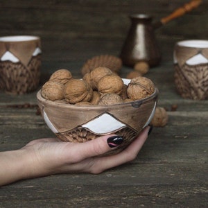 May include: A brown ceramic bowl with white geometric designs filled with walnuts. The bowl is being held by a hand with black nail polish.