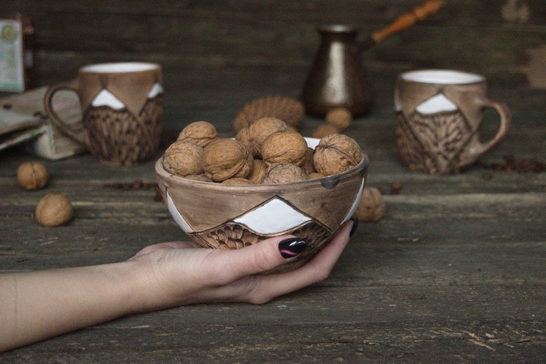 May include: A brown ceramic bowl with white geometric designs filled with walnuts. The bowl is being held by a hand with black nail polish.
