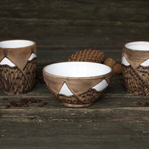 May include: Three ceramic mugs and a bowl with a mountain design. The mugs have brown and white glaze and a handle. The bowl is brown with white accents. The items are on a wooden surface.
