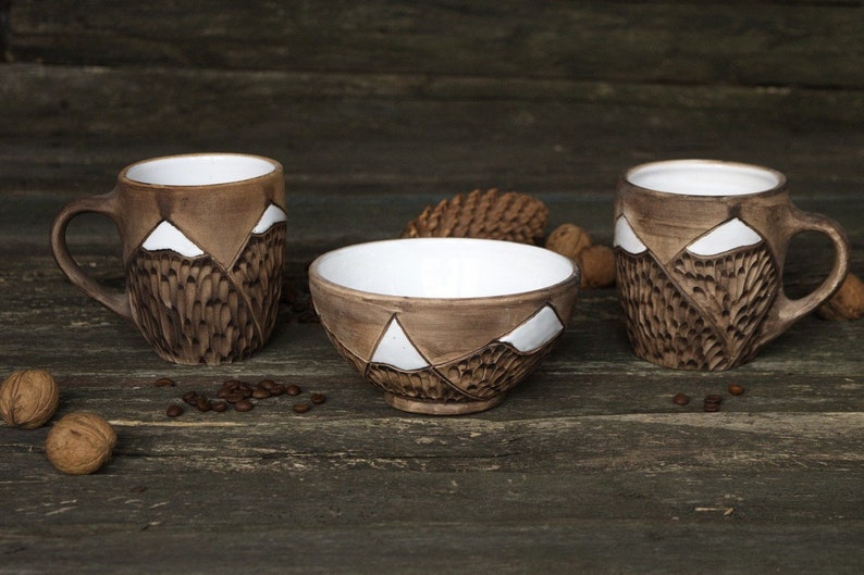 May include: Three ceramic mugs and a bowl with a mountain design. The mugs have brown and white glaze and a handle. The bowl is brown with white accents. The items are on a wooden surface.