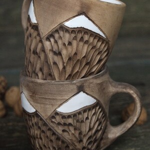 May include: Two brown ceramic mugs with white accents. The mugs have a textured design that resembles feathers or scales. The mugs are stacked on top of each other.
