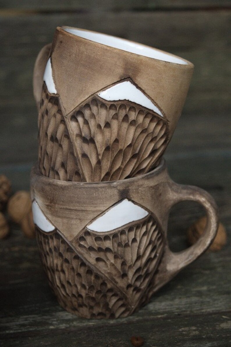 May include: Two brown ceramic mugs with white accents. The mugs have a textured design that resembles feathers or scales. The mugs are stacked on top of each other.