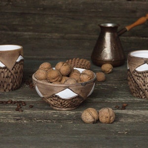 May include: Three brown ceramic mugs with white mountain designs and a brown ceramic bowl filled with walnuts on a wooden surface.