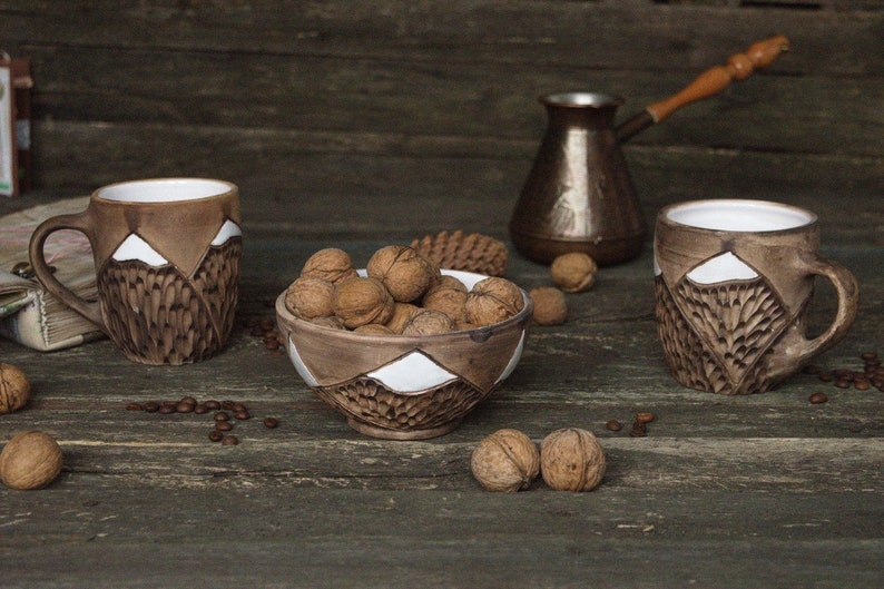 May include: Three brown ceramic mugs with white mountain designs and a brown ceramic bowl filled with walnuts on a wooden surface.