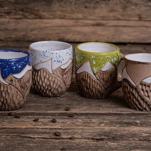 May include: Four ceramic mugs with a mountain design. Each mug has a different colored rim: blue, white, green, and brown. The mugs are sitting on a wooden surface.