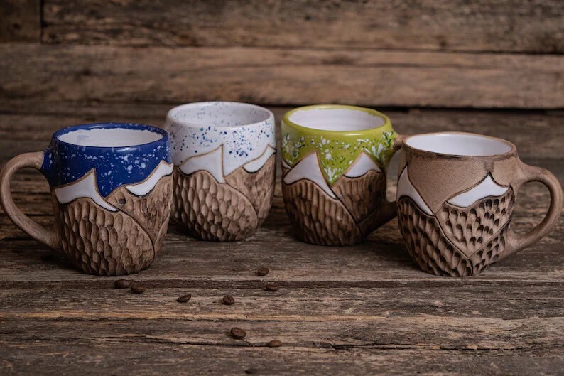 May include: Four ceramic mugs with a mountain design. Each mug has a different colored rim: blue, white, green, and brown. The mugs are sitting on a wooden surface.
