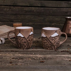 May include: Two brown ceramic mugs with white mountain designs. The mugs are sitting on a wooden surface with coffee beans scattered around them.