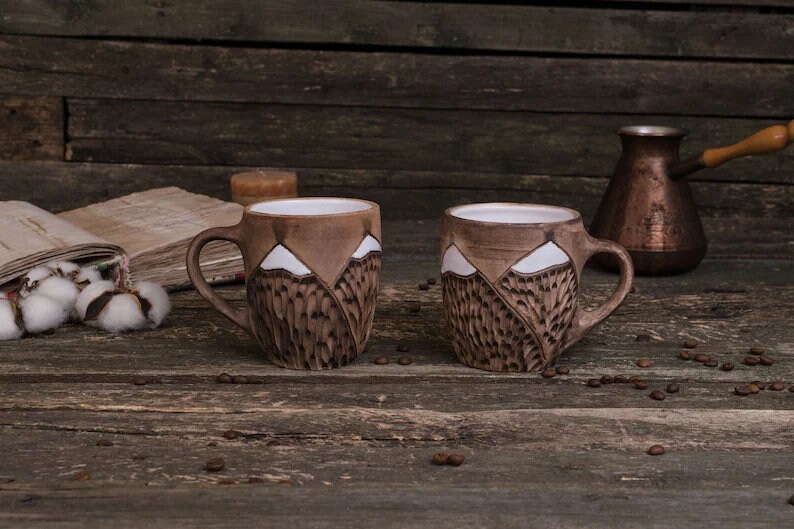 May include: Two brown ceramic mugs with white mountain designs. The mugs are sitting on a wooden surface with coffee beans scattered around them.