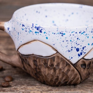 Może przedstawiać: Biały kubek ceramiczny z brązową, fakturowaną podstawą i wzorem w niebieskie i białe plamy. Kubek ma uchwyt i stoi na drewnianej powierzchni.