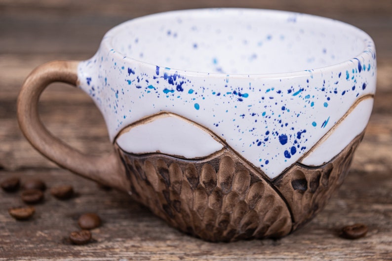 Może przedstawiać: Biały kubek ceramiczny z brązową, fakturowaną podstawą i wzorem w niebieskie i białe plamy. Kubek ma uchwyt i stoi na drewnianej powierzchni.