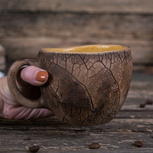 May include: A hand holds a brown ceramic mug with a leaf pattern. The mug has a light brown interior and a wide handle.
