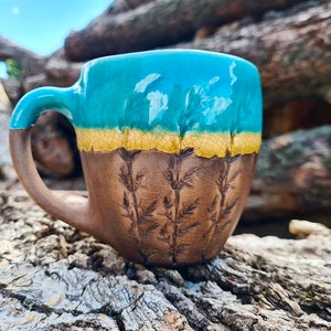 May include: A handmade ceramic mug with a teal glaze and a brown base. The mug has a textured design of leaves and a handle.