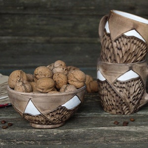 May include: A set of three brown ceramic mugs with white mountain designs and a matching brown ceramic bowl filled with walnuts. The mugs and bowl have a textured surface.