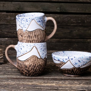 Può includere: Due tazze in ceramica con un design a catena montuosa e macchie blu impilate l'una sull'altra. Una ciotola in ceramica abbinata con un design a catena montuosa e macchie blu si trova davanti alle tazze.