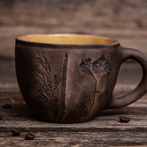 May include: A brown ceramic mug with a yellow interior and a handle. The mug has a rustic look and features an embossed design of plants and flowers.
