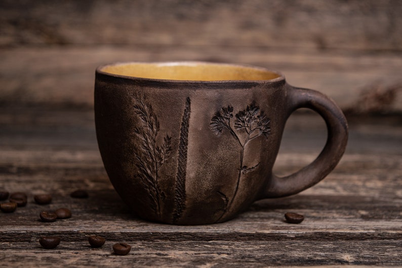 May include: A brown ceramic mug with a yellow interior and a handle. The mug has a rustic look and features an embossed design of plants and flowers.