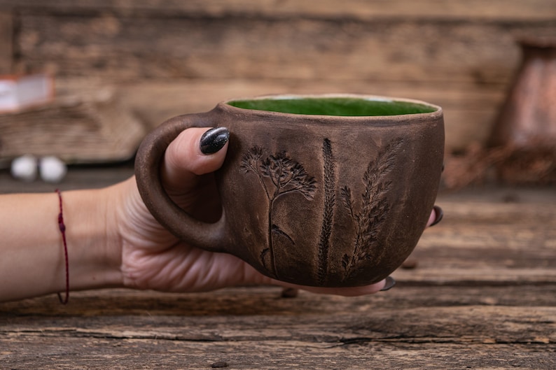 May include: A brown ceramic mug with a green glaze inside and a design of embossed plants on the side. The mug is being held by a hand.