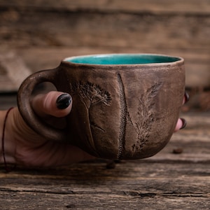 May include: A brown ceramic mug with a teal blue interior and a raised design of grass and flowers. The mug is being held by a hand.