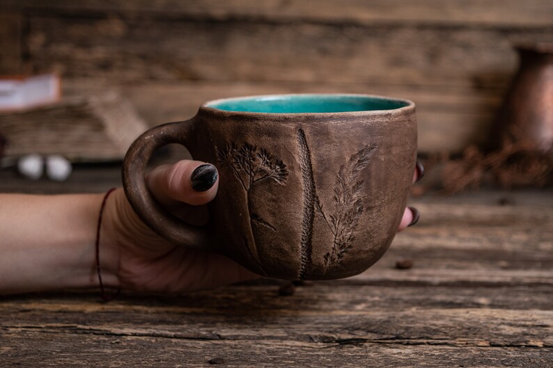 May include: A brown ceramic mug with a teal blue interior and a raised design of grass and flowers. The mug is being held by a hand.