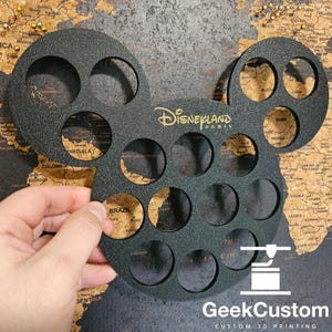 Op de afbeelding: Zwarte Mickey Mouse-vormige wanddecoratie met 18 ronde uitsparingen. De decoratie is gemaakt van een getextureerd materiaal en heeft de woorden "Disneyland Paris" in gouden letters gedrukt.