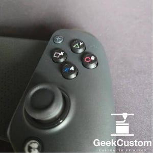 Peut inclure: Une manette de jeu noire avec un joystick circulaire et des boutons portant des symboles : X, O, Triangle, Carré et un pavé directionnel. La manette a une surface texturée et un logo qui dit "GeekCustom CUSTOM 3D PRINTING".