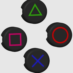 Könnte beinhalten: Vier schwarze Knöpfe mit verschiedenen farbigen Symbolen. Die Knöpfe sind kreisförmig und haben eine kleine Kerbe an der Seite. Die Symbole sind ein grünes Dreieck, ein rosa Quadrat, ein roter Kreis und ein blaues X.