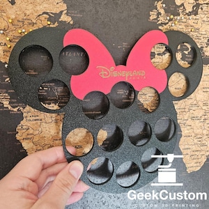 Puede incluir: Un soporte para corchos impreso en 3D en forma de cabeza de Mickey Mouse en negro y rojo con el texto "Disneyland Paris" en letras doradas. El soporte tiene 16 agujeros para corchos.