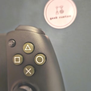 May include: A black video game controller with a circular button layout. The buttons are labeled with white symbols: square, triangle, circle, and X. The controller has a black joystick and a gray trigger.