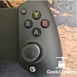 May include: A black gaming controller with a textured surface. The controller has a circular button with a white "A" on it, a circular button with a red "B" on it, a circular button with a yellow "Y" on it, and a circular button with a blue "X" on it. The controller also has a black joystick and a black button with a plus sign on it. The controller is labeled "GeekCustom CUSTOM 3D PRINTING".