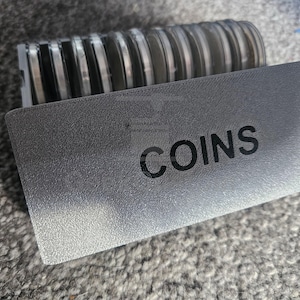 Può includere: Una scatola portaoggetti grigia stampata in 3D con una superficie ruvida e la parola COINS stampata in nero sul davanti. La scatola contiene una fila di diverse monete circolari, ciascuna racchiusa singolarmente in una capsula protettiva.