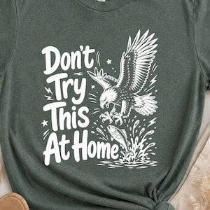 Könnte beinhalten: Ein dunkelwaldgrünes Kurzarm-T-Shirt mit einem weißen Grafikdruck eines Weißkopfseeadlers, der einen Fisch fängt. Der Text "Don't Try This At Home" ist links neben der Adlerillustration in einer fetten, weißen Schrift aufgedruckt.