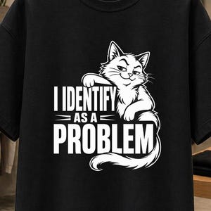 May include: A black short-sleeved t-shirt featuring a white graphic of a cat leaning over the text that reads I IDENTIFY AS A PROBLEM in bold capital letters. The design is centered on the chest of the garment, which is displayed on a wooden hanger.