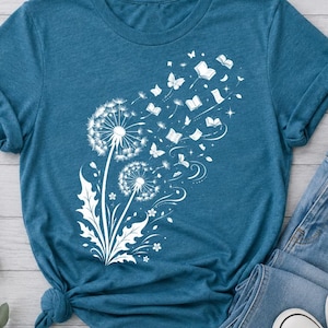 Peut inclure: Un t-shirt bleu sarcelle à manches courtes avec une impression graphique blanche représentant deux pissenlits. Les graines des pissenlits se transforment en petits livres volants et en papillons, créant une traînée sur le devant du vêtement.