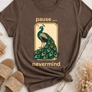 May include: A dark brown short-sleeved t-shirt featuring a graphic of a peacock with a large tail. Above the peacock is the text "pause ..." and below it is the text "nevermind". The graphic is enclosed in a rectangular border with a light background.