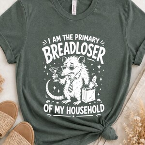 May include: A dark green t-shirt featuring a white graphic of an opossum wearing sunglasses and a necktie, holding a martini glass and a paper bag of bread. The text reads I AM THE PRIMARY BREADLOSER OF MY HOUSEHOLD in a bold, arched font.