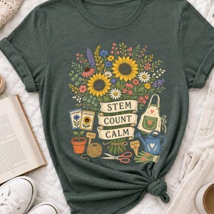 Peut inclure: Un t-shirt vert foncé avec un motif coloré représentant un bouquet de tournesols et de fleurs sauvages. Une bannière centrale affiche le texte "STEM COUNT CALM" entouré d'illustrations d'outils de jardinage, de sachets de graines et d'un pot.